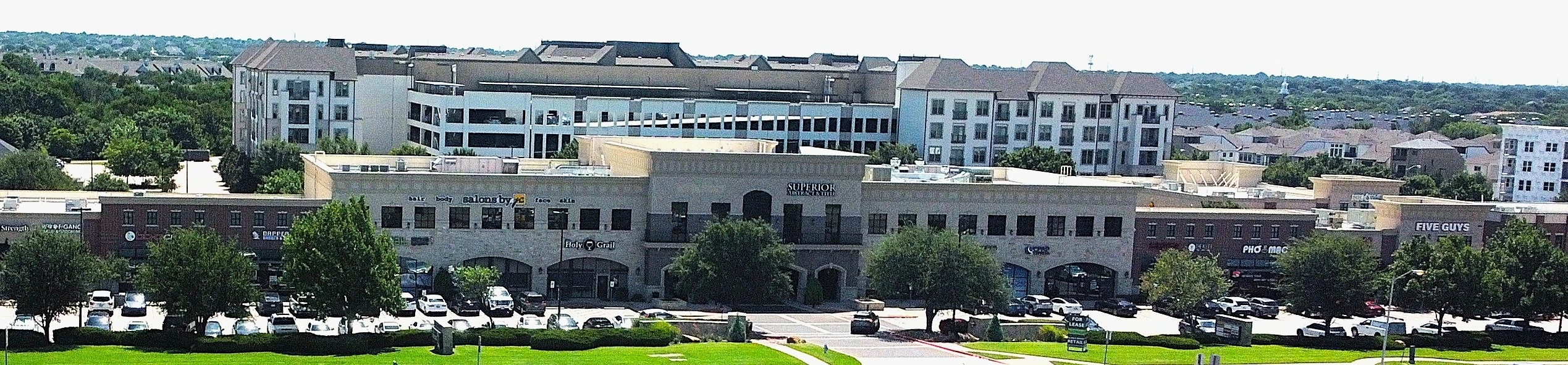 Wide aerial view of Headquarters Village Shopping Center's retail storefronts with luxury apartments in the background, Plano, Texas